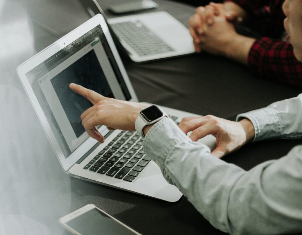 A man sat at a desk with a colleague, pointing at his laptop screen